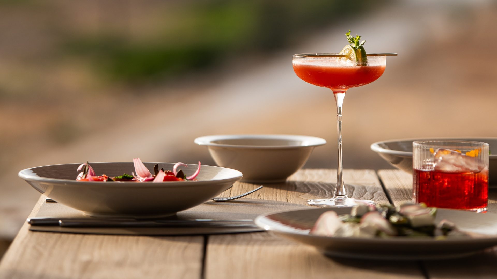 A coupe of red cocktail garnished with lime and mint beside small plates on a bare wooden table