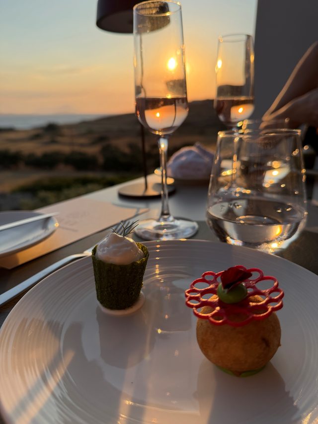 The two amuse-bouche, spanakopita and tomato fritter, on a white plate at a terrace table with the sun setting over the sea behind