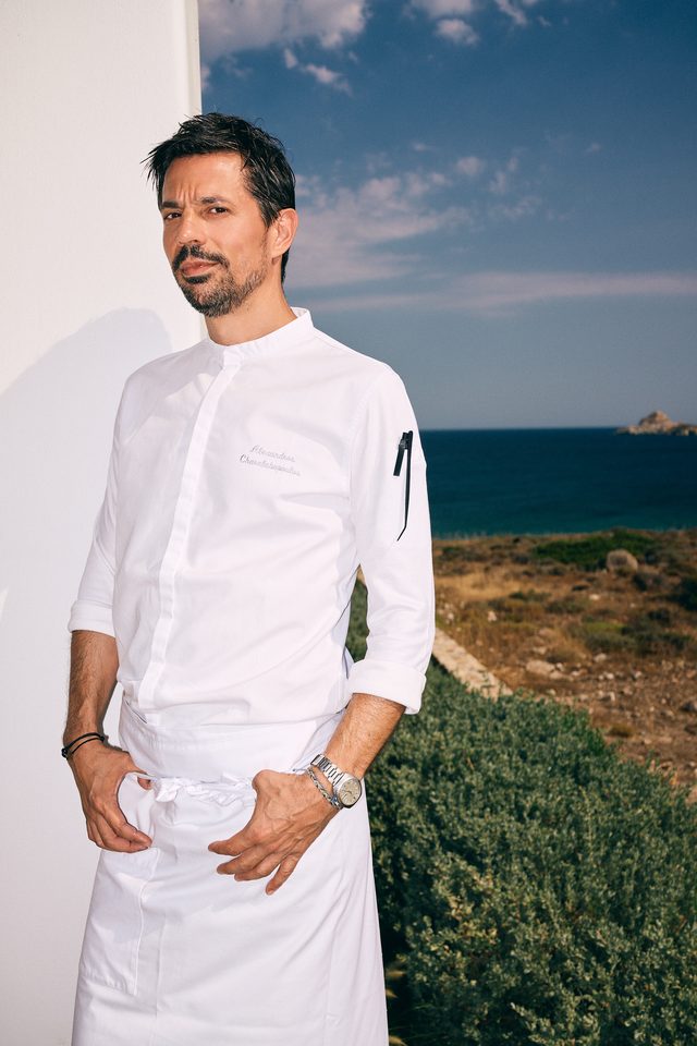 Guest chef Alexandros Charalabopoulos in his whites, standing before the low scrub of Kimolos and the open Aegean