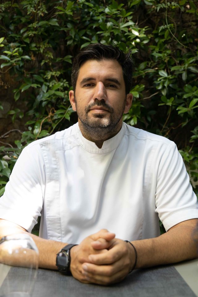 Guest chef Ghikas Xenakis in his whites, seated at a table with his hands clasped, dense green foliage behind him