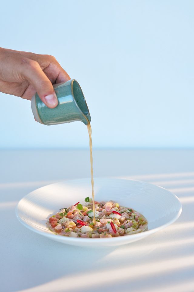 A hand tipping a small green jug of broth over a shallow bowl of diced raw fish, chilli and herbs, against a pale blue sky