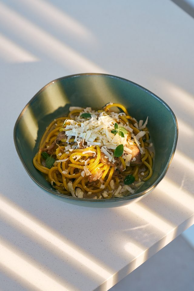 Golden fresh spaghetti with slow-cooked meat under a heap of grated dry cheese, in a green ceramic bowl on a sunlit ledge