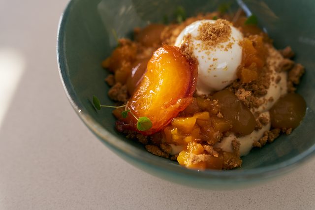 Roasted peach wedges with a quenelle of ice cream, poached grapes and buttery crumble, in a green ceramic bowl