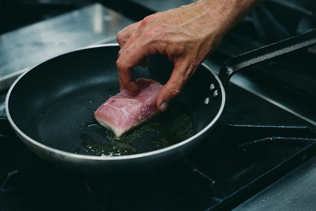 A cook's hand laying a fillet of fish into hot oil in a black pan