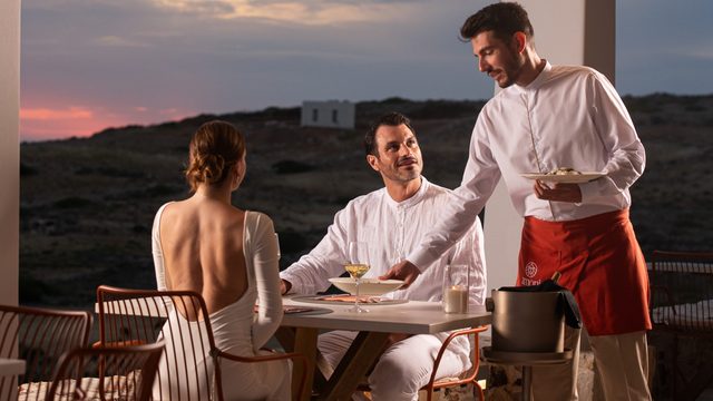A waiter in an amoni apron setting a plate down for two guests at a table outdoors, the sky pink behind the dry hills