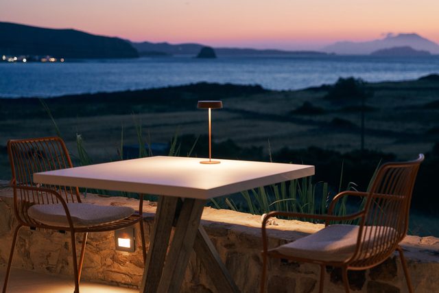 A single table lit by a small lamp at dusk, the sky pink over the dark sea and the outline of Milos