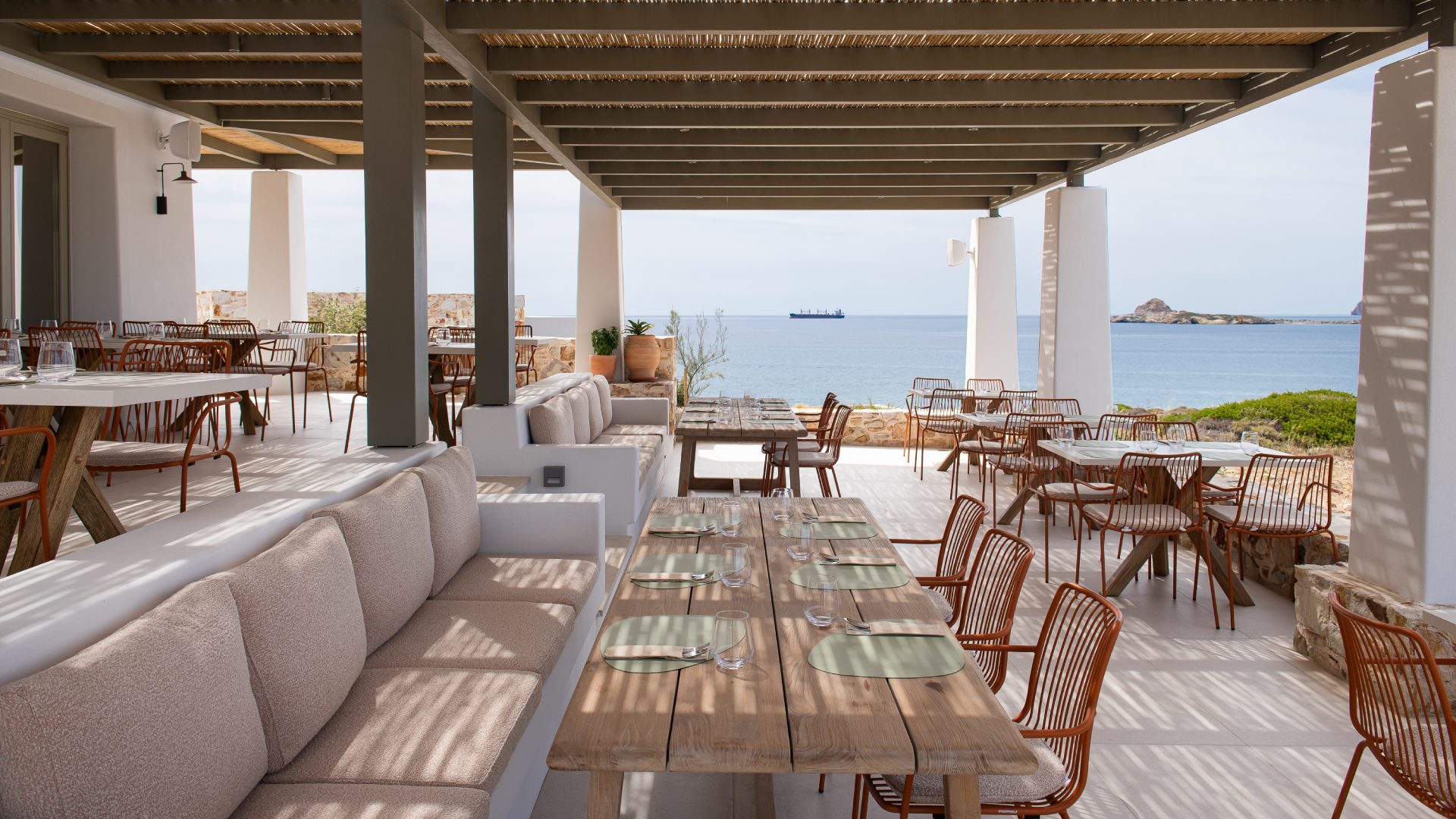 The terrace by day, striped shadow from the reed pergola falling across long wooden tables that look straight out over the Aegean
