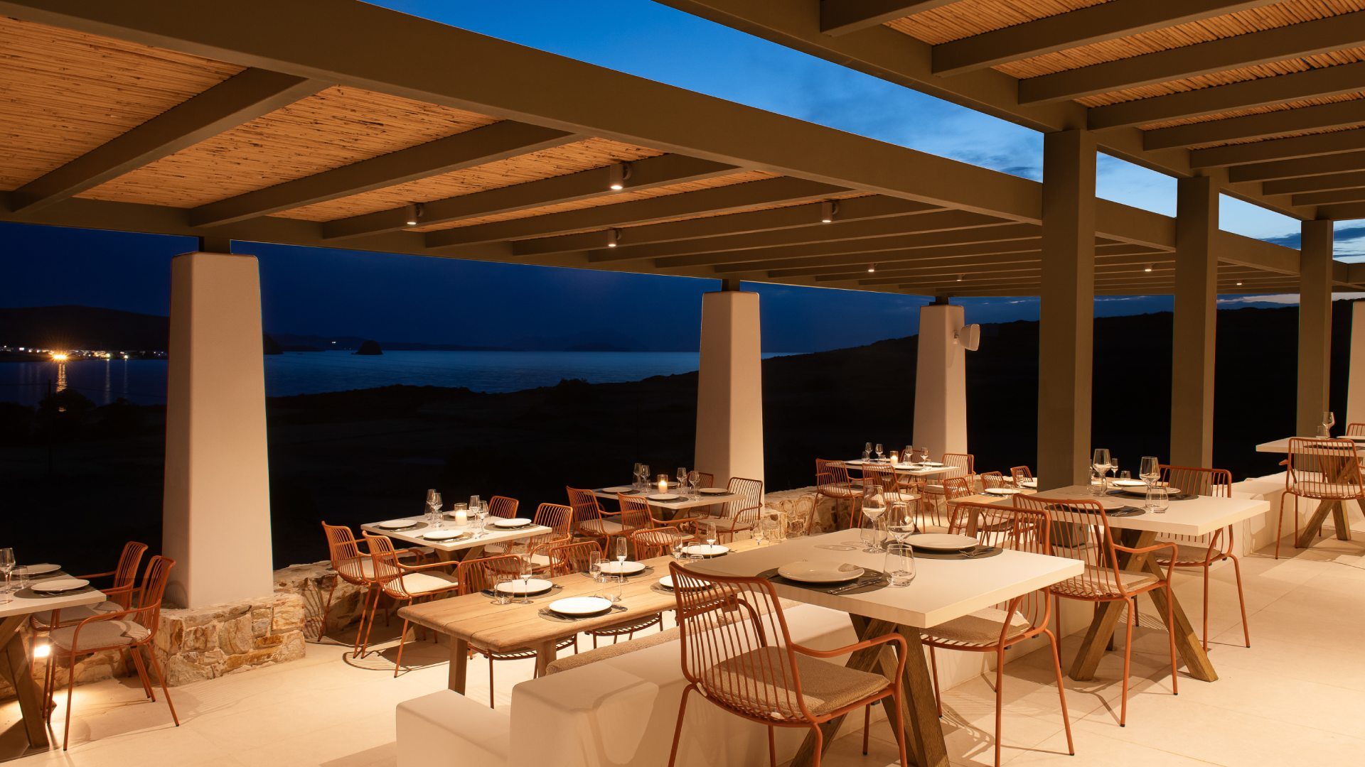 Amoni's open-air terrace at dusk, tables set under a reed pergola with terracotta chairs, the dark sea and the lights of Kimolos beyond