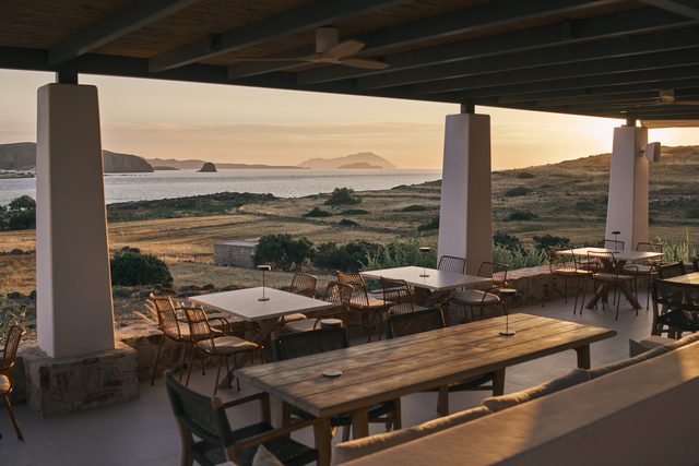 The terrace at sunset, long wooden tables and terracotta chairs under the pergola, looking across dry ground to the sea and distant islands