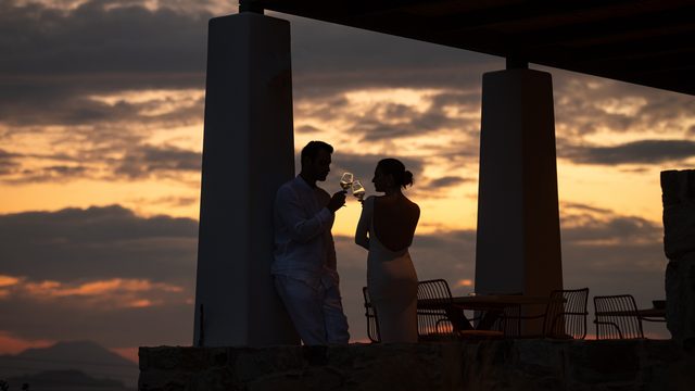 Two people silhouetted between white columns, touching wine glasses against an orange and grey sunset